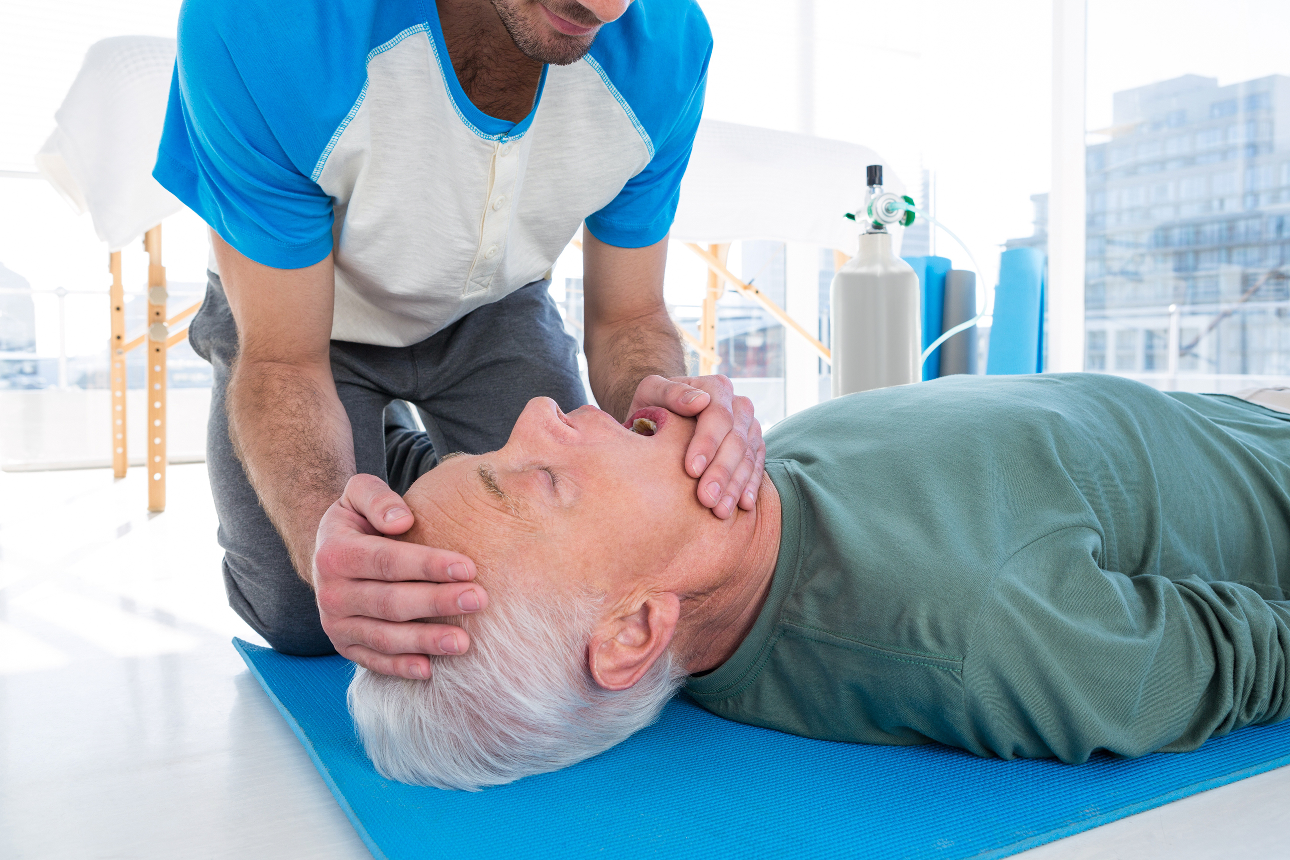 Patient on floor about to receive CPR from man kneeling at his side with emergency oxygen cylinder in the background