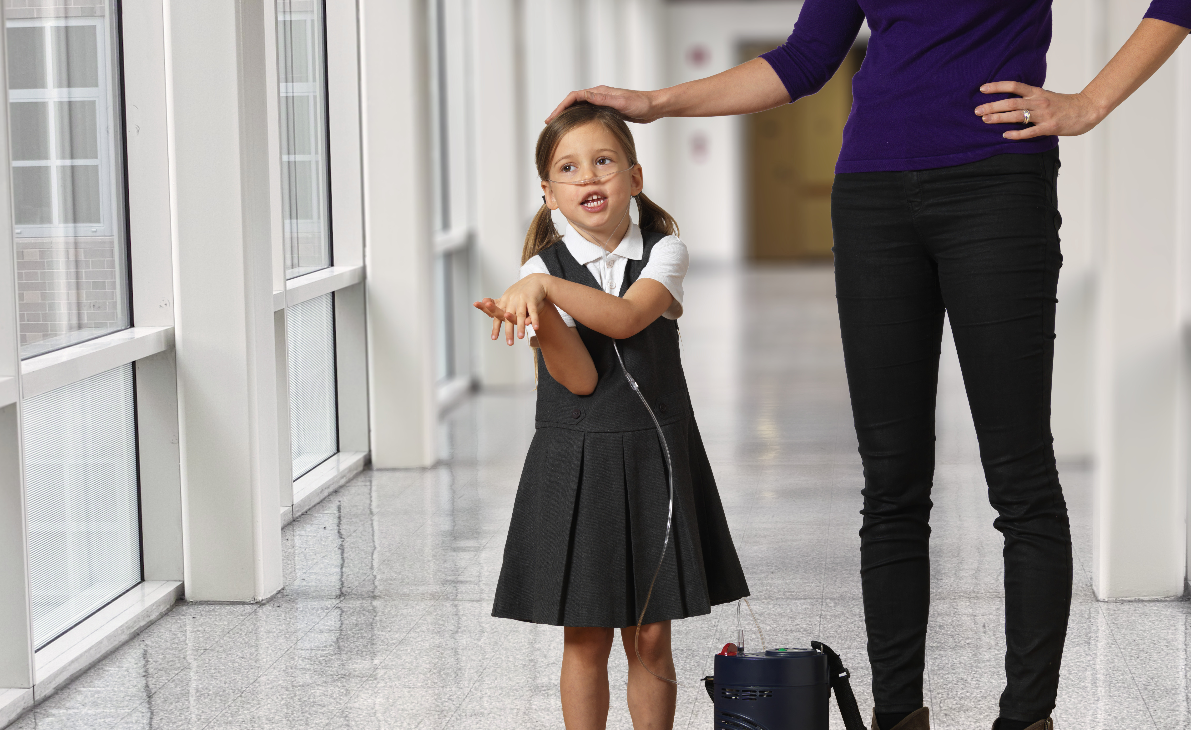 little girl patient with portable oxygen concentrator stood with parent in school corridor